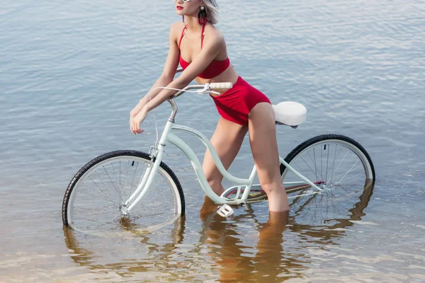 Cropped view of slim girl in red trendy bikini posing with bicycle in water — Stock Photo