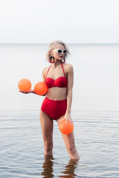 Fille à la mode en bikini rouge vintage et lunettes de soleil posant dans l'eau avec des boules orange — Photo de stock