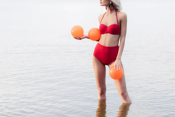 Ausgeschnittene Ansicht eines stilvollen Mädchens im roten Bikini, das im Wasser steht und orangefarbene Kugeln in der Hand hält — Stockfoto