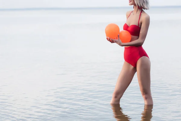 Corte vista de mulher em biquíni vermelho em pé na água e segurando bolas laranja — Fotografia de Stock