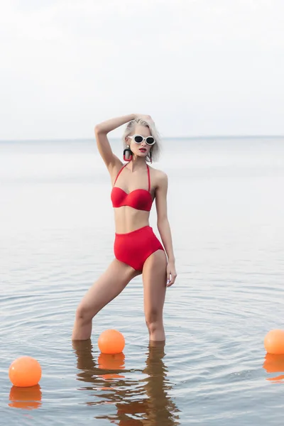 Hermosa mujer en bikini rojo retro y gafas de sol posando en agua con bolas - foto de stock