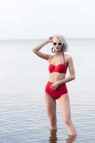 Atractiva mujer de moda en bikini rojo vintage y gafas de sol de pie en el agua en el mar - foto de stock