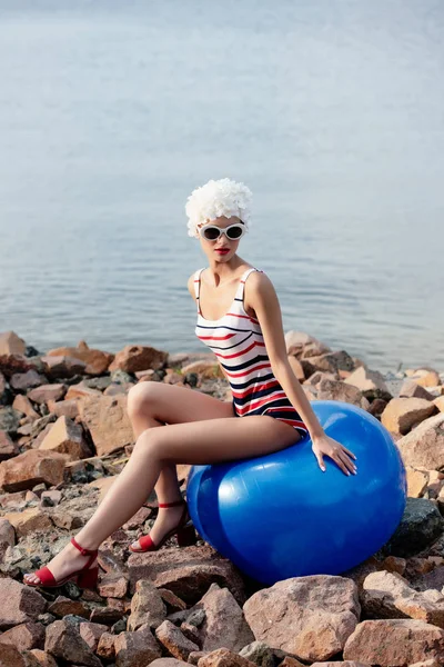 Стильна приваблива дівчина в старовинному смугастому купальнику і сонцезахисних окулярах, що сидять на синьому м'ячі на кам'янистому пляжі — стокове фото