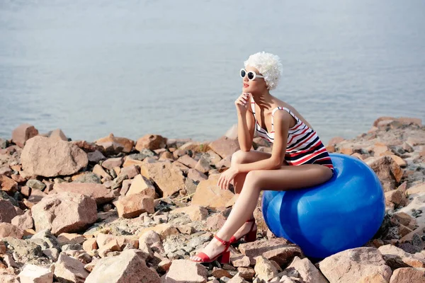 Stylish girl in retro striped swimsuit sitting on blue fitness ball on rocky beach — Stock Photo