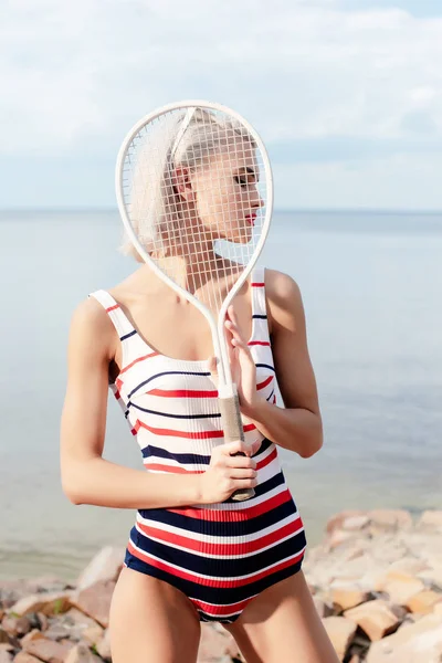 Блондинка приваблива жінка в смугастому купальнику позує з тенісною ракеткою на березі — стокове фото