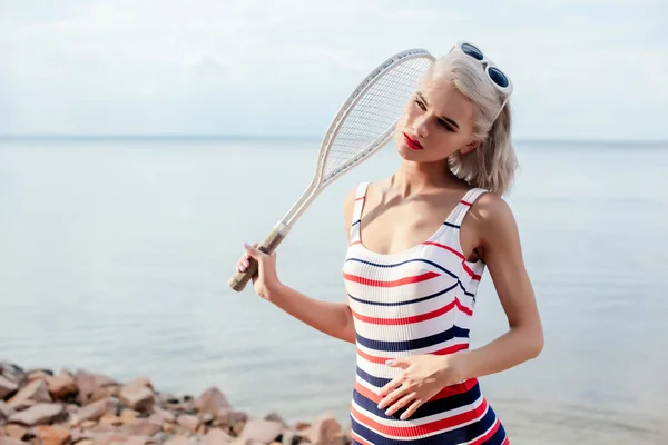 Schöne blonde Sportlerin im gestreiften Badeanzug posiert mit Tennisschläger am Meer — Stockfoto
