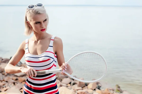Atractiva chica rubia en traje de baño a rayas y gafas de sol posando con raqueta de tenis en la playa en el mar - foto de stock