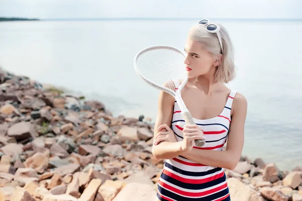 Элегантная девушка в ретро-полосатых купальниках позирует с теннисной ракеткой на скалистом пляже — стоковое фото