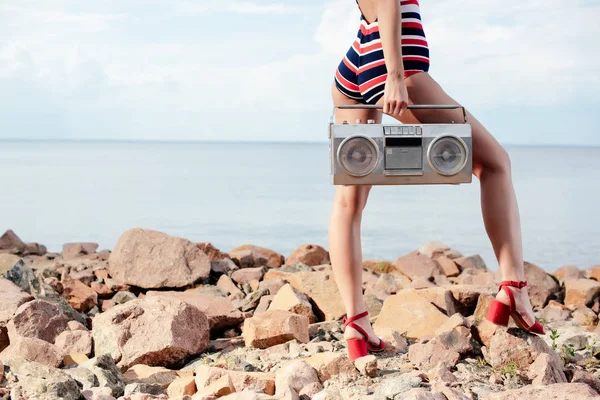 Vista ritagliata della donna in costume da bagno in possesso di boombox vintage su rocce vicino al mare — Foto stock