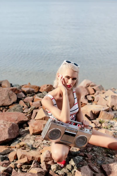 Beautiful blonde woman in swimsuit posing with vintage boombox on rocky beach — Stock Photo
