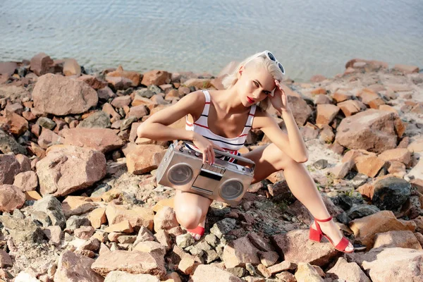 Beautiful fashionable girl posing with retro boombox on rocky beach — Stock Photo