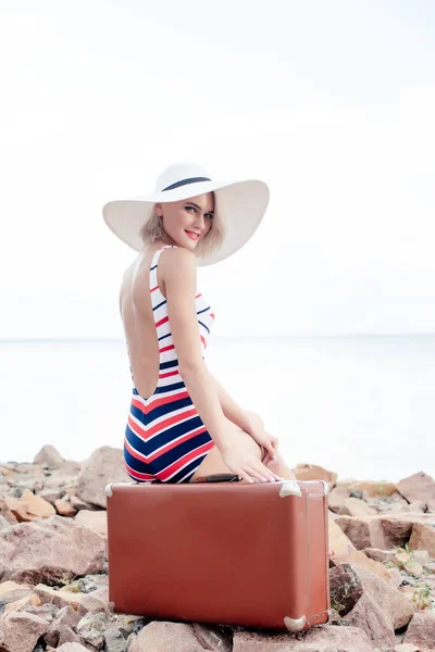 Female tourist in swimsuit and white hat sitting on vintage travel bag on rocky beach — Stock Photo