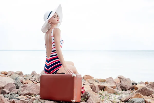 Beautiful woman in striped swimsuit and hat sitting on retro travel bag on rocky beach — Stock Photo
