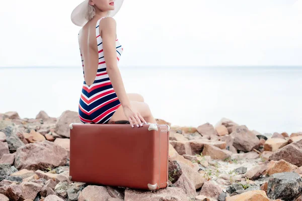 Cropped view of stylish woman in striped swimsuit sitting on luggage on rocky beach — Stock Photo