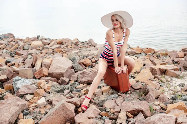Hermosa turista femenina en traje de baño sentado en la bolsa de viaje vintage en la playa rocosa - foto de stock