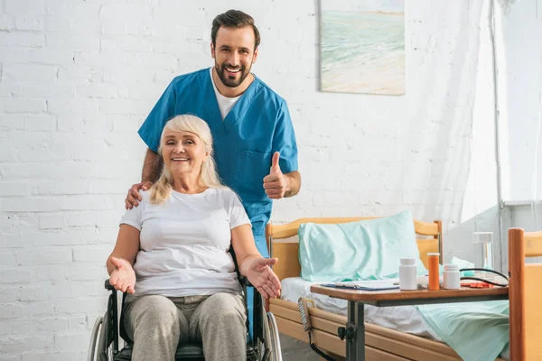 Feliz anciana en silla de ruedas sonriendo a la cámara y alegre joven médico mostrando el pulgar hacia arriba - foto de stock