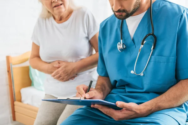 Recortado tiro de doctor escribiendo en portapapeles mientras enfermo senior mujer sentado en hospital cama - foto de stock