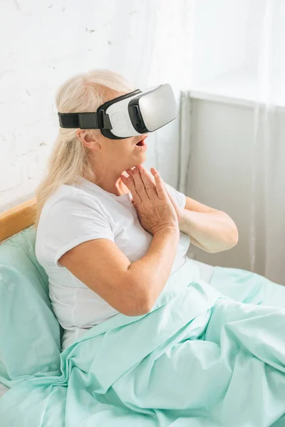 Высокий угол обзора шокированной пожилой женщины с помощью гарнитуры виртуальной реальности на больничной койке — стоковое фото