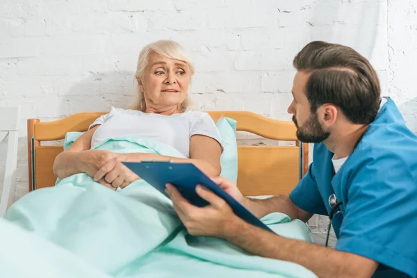 Infirmière masculine écrivant sur presse-papiers et regardant malade femme âgée couché dans le lit d'hôpital — Photo de stock