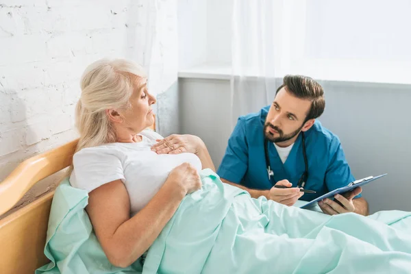 Médecin écrit sur presse-papiers et regardant la femme âgée malade couché dans le lit d'hôpital — Photo de stock