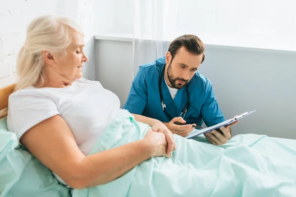 Doctor writing on clipboard while sick senior woman lying in hospital bed — Stock Photo
