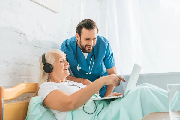Enfermera masculina sonriente mirando a la mujer mayor feliz en los auriculares usando el ordenador portátil en la cama del hospital - foto de stock