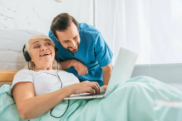 Lächelnder Arzt blickt glückliche Seniorin mit Kopfhörer an und benutzt Laptop im Krankenhausbett — Stockfoto