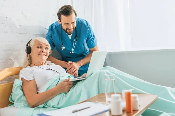 Улыбающийся молодой врач смотрит на счастливую пожилую женщину в наушниках с помощью ноутбука в постели — стоковое фото