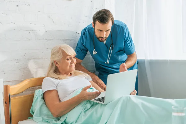 Doctor with stethoscope looking at senior woman using laptop in hospital bed — Stock Photo