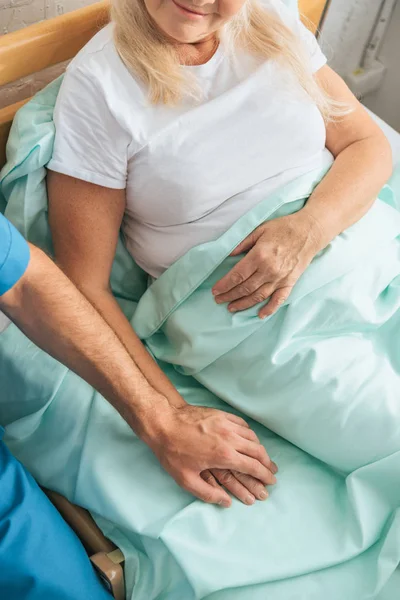 Vue partielle de l'infirmier tenant la main d'une femme âgée malade couchée dans un lit d'hôpital — Photo de stock