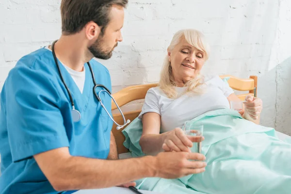 Young doctor with stethoscope giving glass of water to senior woman lying in hospital bed — Stock Photo
