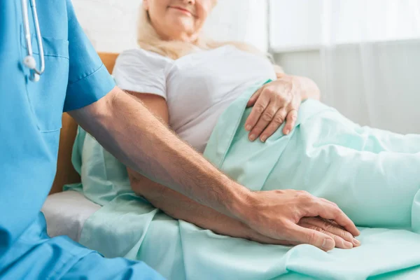 Tiro recortado de la enfermera masculina cogida de la mano de la mujer anciana enferma acostada en la cama del hospital - foto de stock