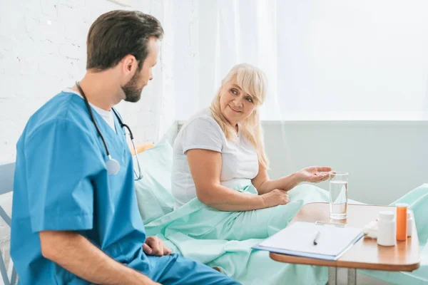 Arzt mit Stethoskop betrachtet Seniorin, die Medikamente im Krankenhausbett einnimmt — Stockfoto