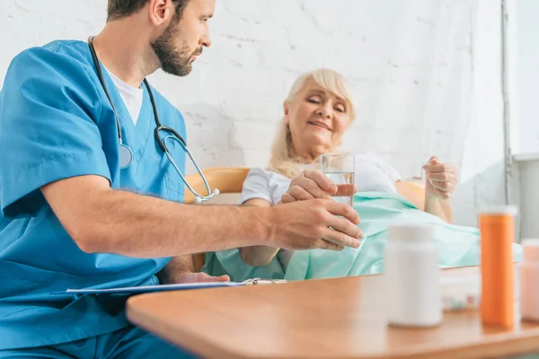 Schnappschuss von junger Ärztin, die Seniorin, die Medikamente im Bett nimmt, ein Glas Wasser gibt — Stockfoto