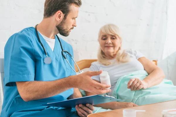 Arzt hält Klemmbrett und zeigt im Bett liegende Seniorin Tabletten — Stockfoto