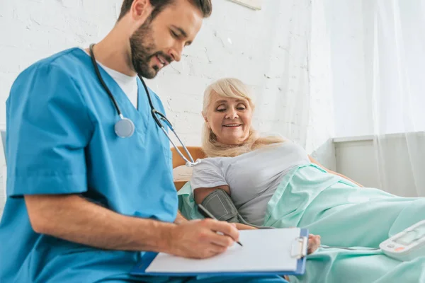 Enfermera masculina sonriente escribiendo en el portapapeles midiendo la presión arterial a una mujer mayor acostada en la cama - foto de stock