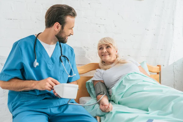 Enfermera sonriente con estetoscopio que sostiene el monitor de presión arterial mientras mide la presión arterial a una mujer mayor acostada en la cama - foto de stock