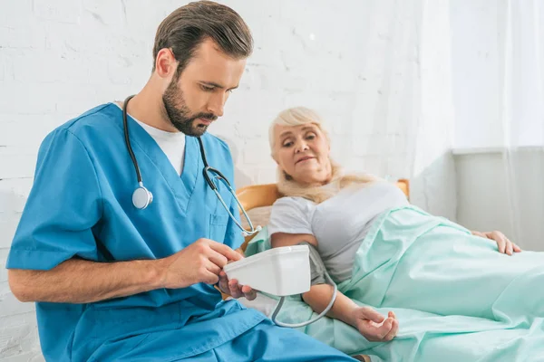 Junge männliche Krankenschwester mit Stethoskop hält Blutdruckmessgerät in der Hand, während sie den Blutdruck der im Bett liegenden Seniorin misst — Stockfoto