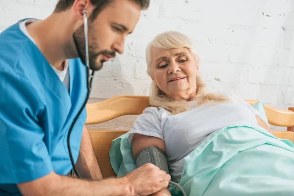 Молодой медбрат измеряет артериальное давление пожилой женщине на больничной койке — стоковое фото