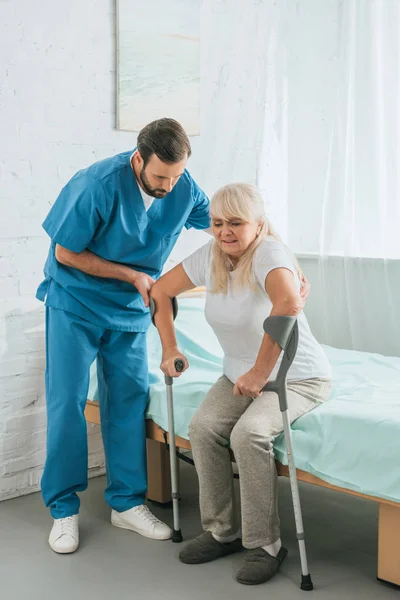 Мужчина медсестра помогает пожилой женщине с костылями, сидя на больничной койке — стоковое фото