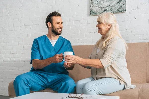 Smiling male nurse giving cup with hot beverage to happy senior woman — Stock Photo