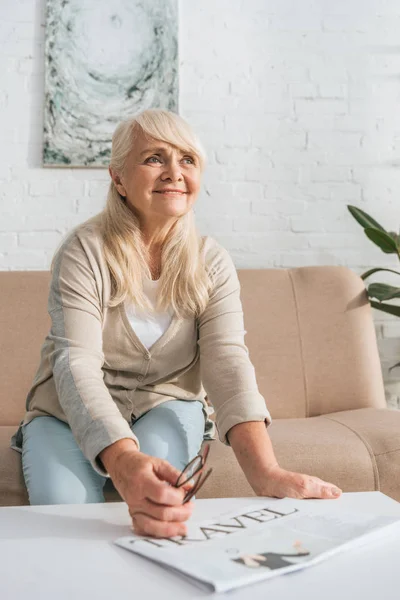 Mujer mayor sonriente sosteniendo anteojos y mirando hacia otro lado mientras está sentada en el sofá en casa - foto de stock