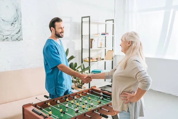 High angle view of smiling senior woman and caregiver shaking hands above table football — Stock Photo