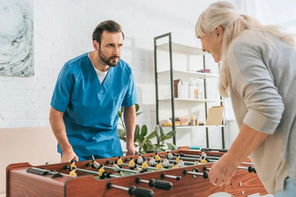 Young male social worker playing table football with smiling senior woman — Stock Photo