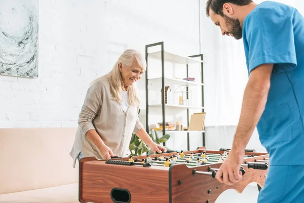 Smiling senior woman and young social worker playing table football together — Stock Photo