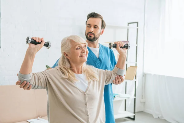 Social worker looking at smiling senior woman training with dumbbells — Stock Photo