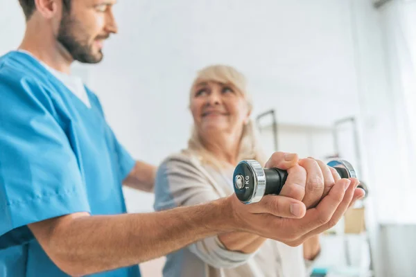Close-up view of social worker helping smiling senior woman exercising with dumbbells — Stock Photo