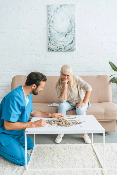 Smiling senior woman sitting with hand on chin and looking at young social worker playing with jigsaw puzzle — Stock Photo