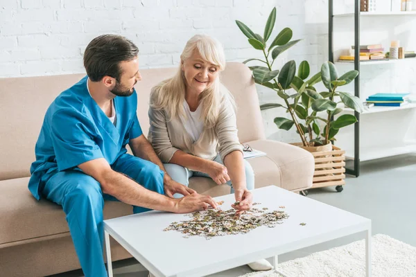 Caregiver looking at smiling senior woman playing with jigsaw puzzle pieces — Stock Photo