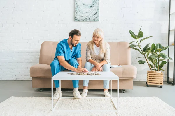 Smiling senior woman and young social worker playing with jigsaw puzzle — Stock Photo